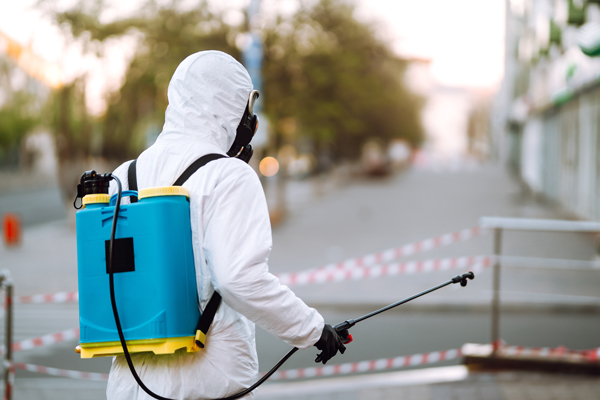 A person wearing a white protective suit, equipped with a backpack and sprayer, stands ready for a cleaning or spraying task.