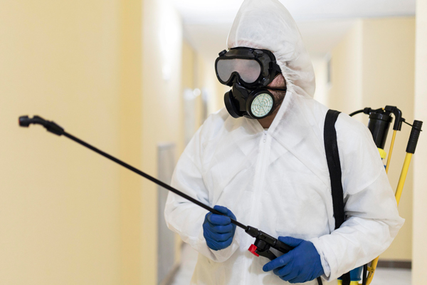 A man in a white protective suit and mask is holding a sprayer, ready to apply a substance for safety or sanitation purposes.